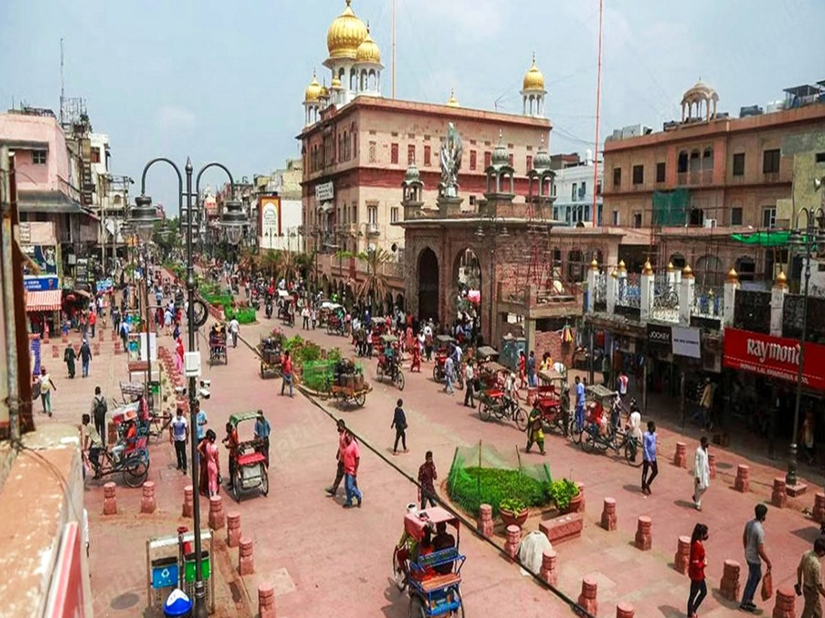 Crowded Chandni Chowk lane lined with brightly lit shops selling spices, fabrics, and jewelry, with shoppers and rickshaws weaving through.