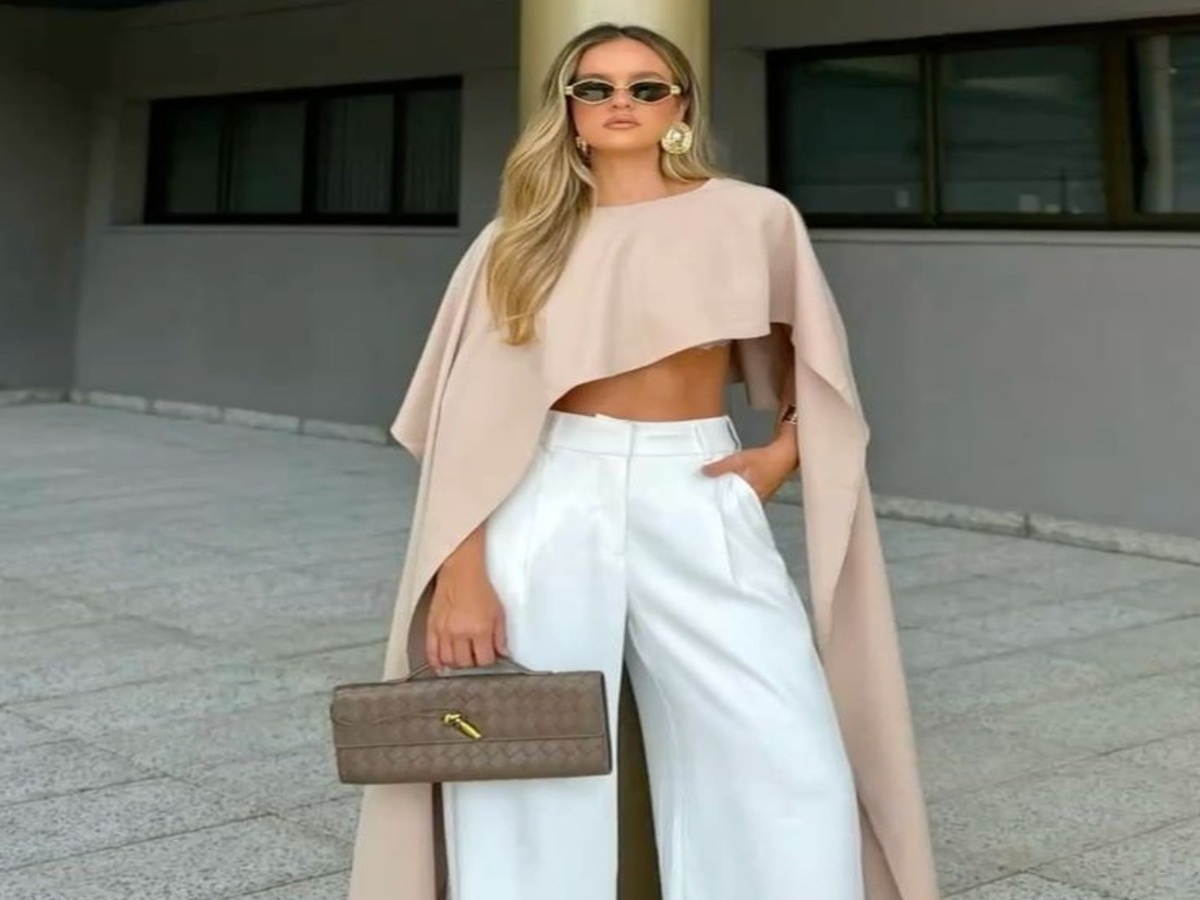 Model wearing a beige cape top and wide white trousers, holding a stylish handbag, set against a minimalist architectural background.