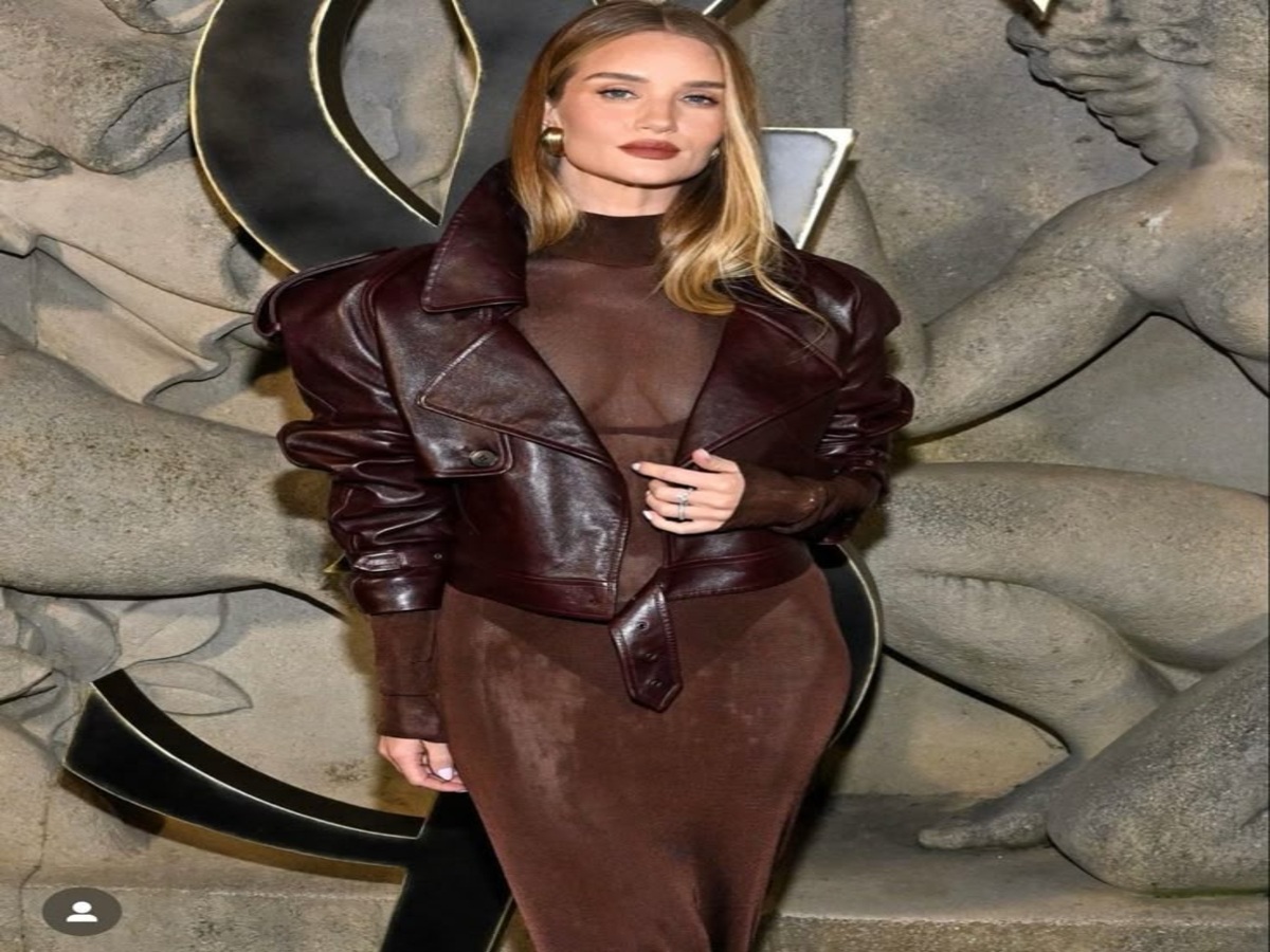 Model in a burgundy leather jacket over a sheer bodysuit, posing against a textured backdrop.