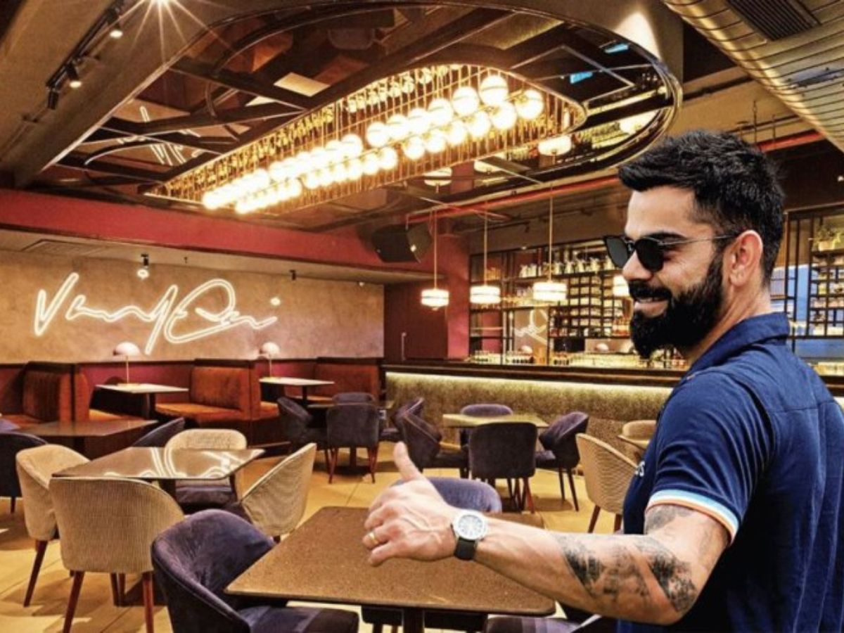 Sunlit glass-roof dining area with rattan hanging lamps, artistic sculptures, and pastel-toned seating in a European-style cafe.