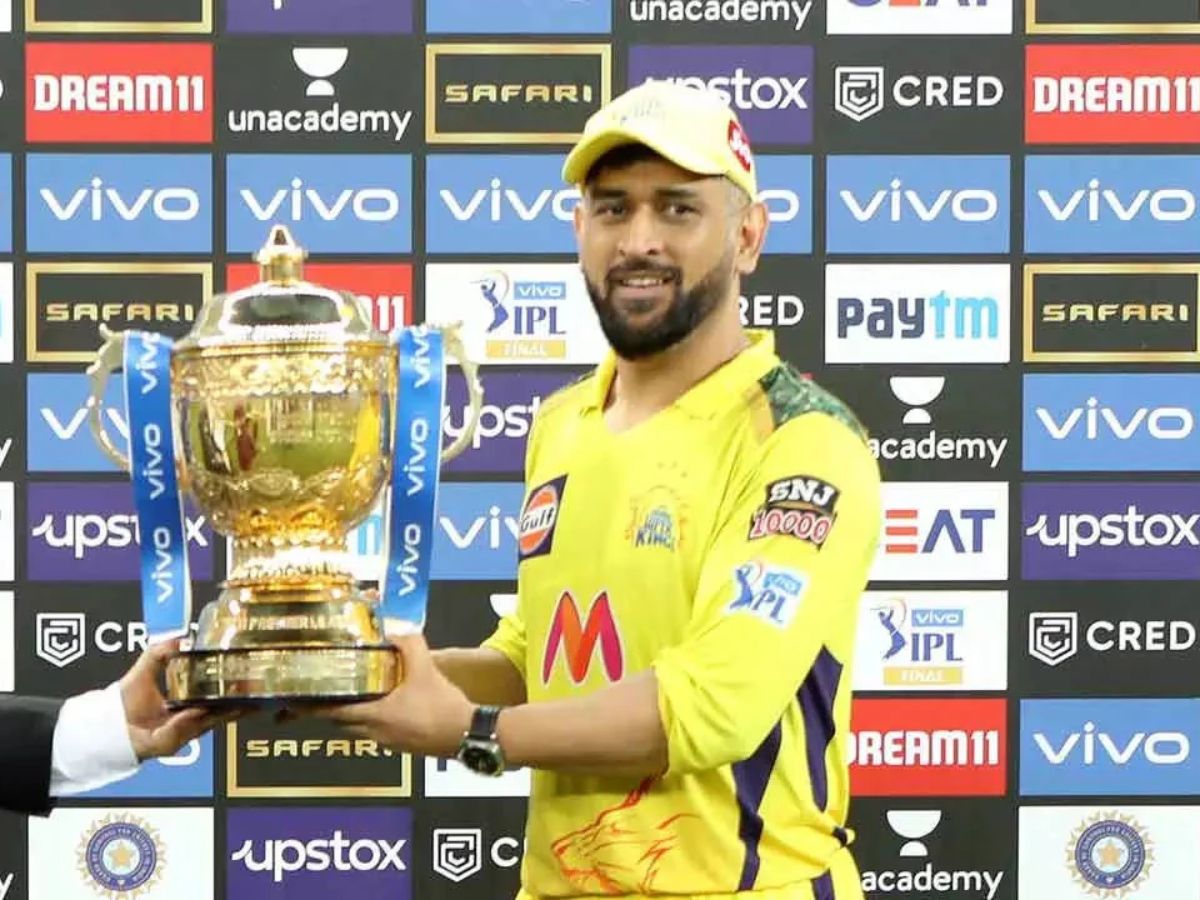 MS Dhoni holding IPL 2023 trophy with wide smile and fans cheering.