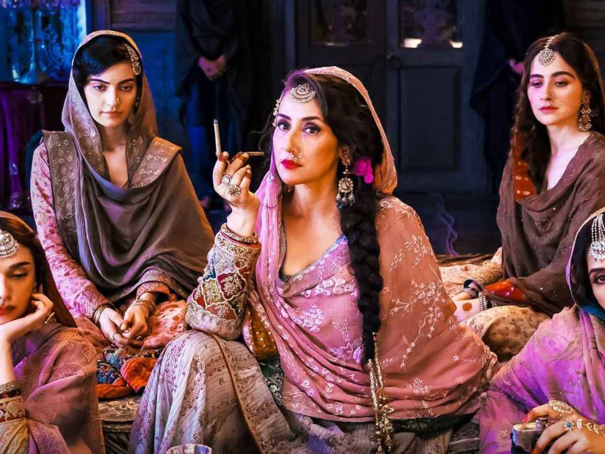 Manisha Koirala as Mallikajaan, sitting and holding a cigarette, wearing a dusty pink and gold gharara and a long braided hairstyle with a paranda. She is surrounded by other women in traditional attire.