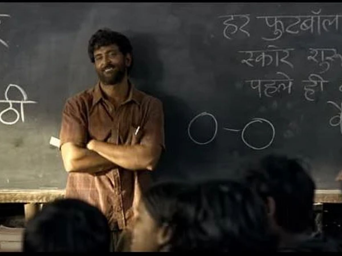 Hrithik Roshan as Anand Kumar standing in front of a blackboard with folded arms, smiling while teaching poor students in a classroom scene from Super 30.