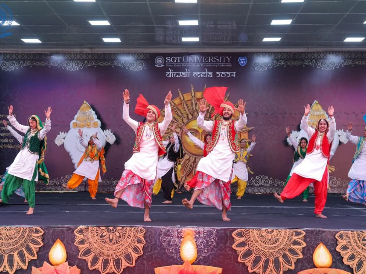 A group of male and female dancers in traditional Indian attire perform on a stage during Diwali celebrations at SGT University. The stage is decorated with large sun and diya motifs.