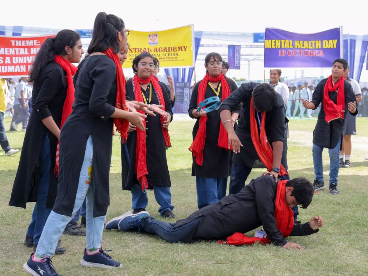Students in black shirts and red scarves perform a skit on mental health awareness in a field. One student is on the ground, and a banner says 'Mental Health Day'.