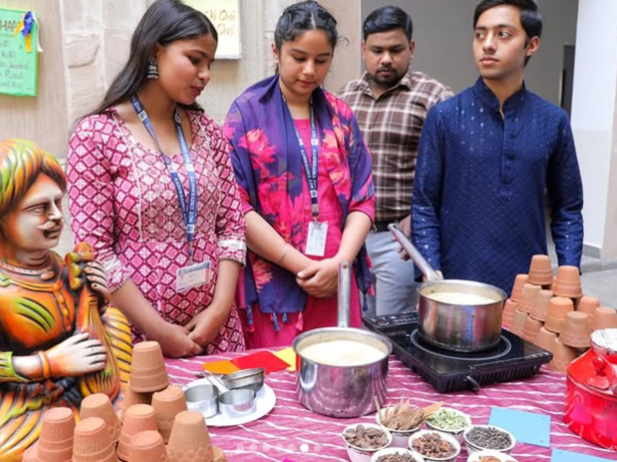 Three students and a man in a plaid shirt stand around a table with two pots of tea on a burner, surrounded by spices and clay cups, at a college event.