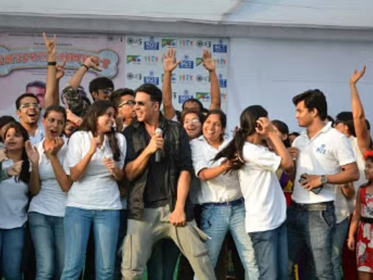 A crowd of students in white shirts and jeans cheering and raising their hands. Actor Akshay Kumar stands in the middle, holding a microphone and smiling.