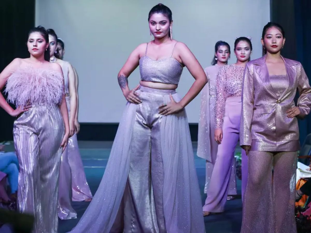 A group of models on a runway. The central model in the foreground wears a shimmering silver-lavender crop top and pants with a sheer skirt overlay.
