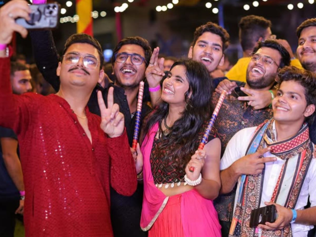 A group of young adults smiling and taking a selfie at a Navratri event. They are holding dandiya sticks and are dressed in festive Indian clothing.