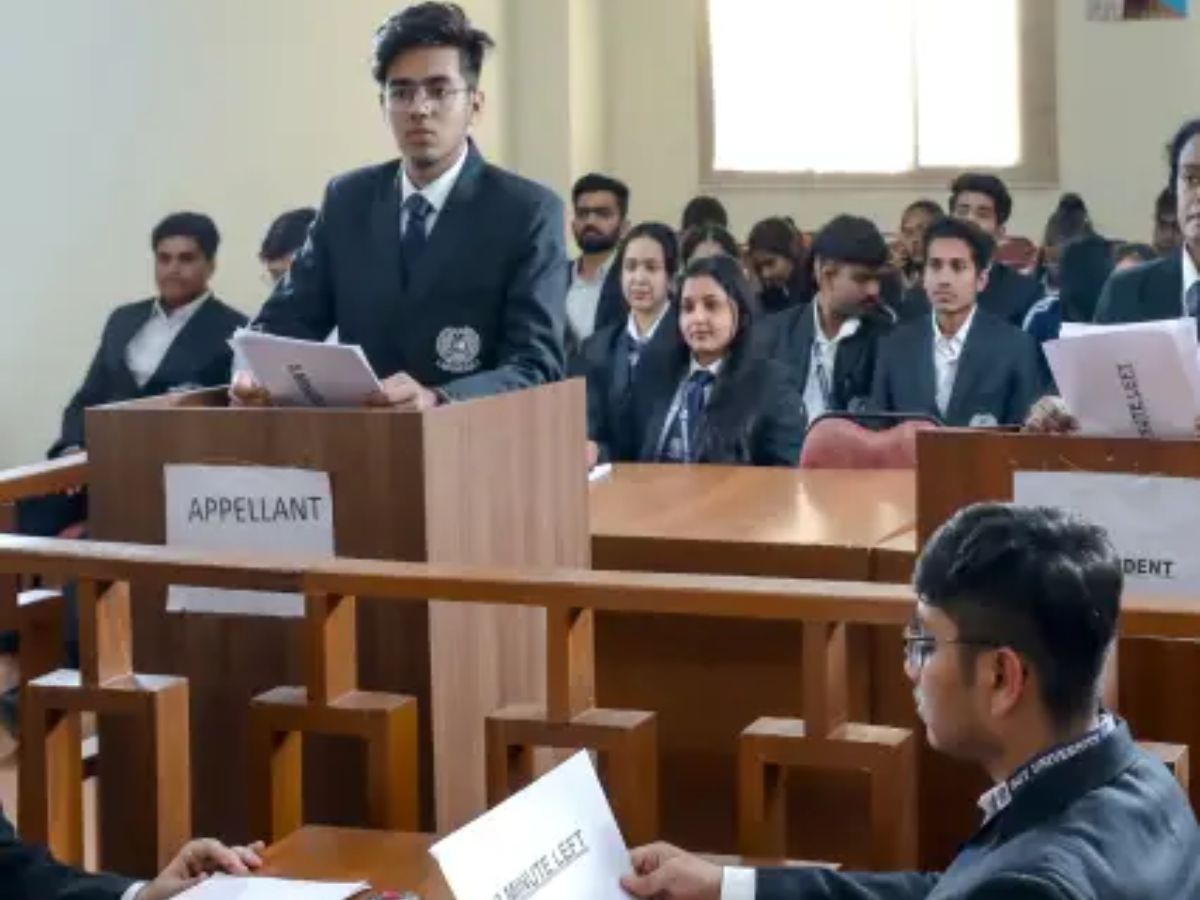 A male student in a black suit stands at a podium marked 'APPELLANT' and speaks, holding a paper. Other students are seated in the background in a mock courtroom setting.