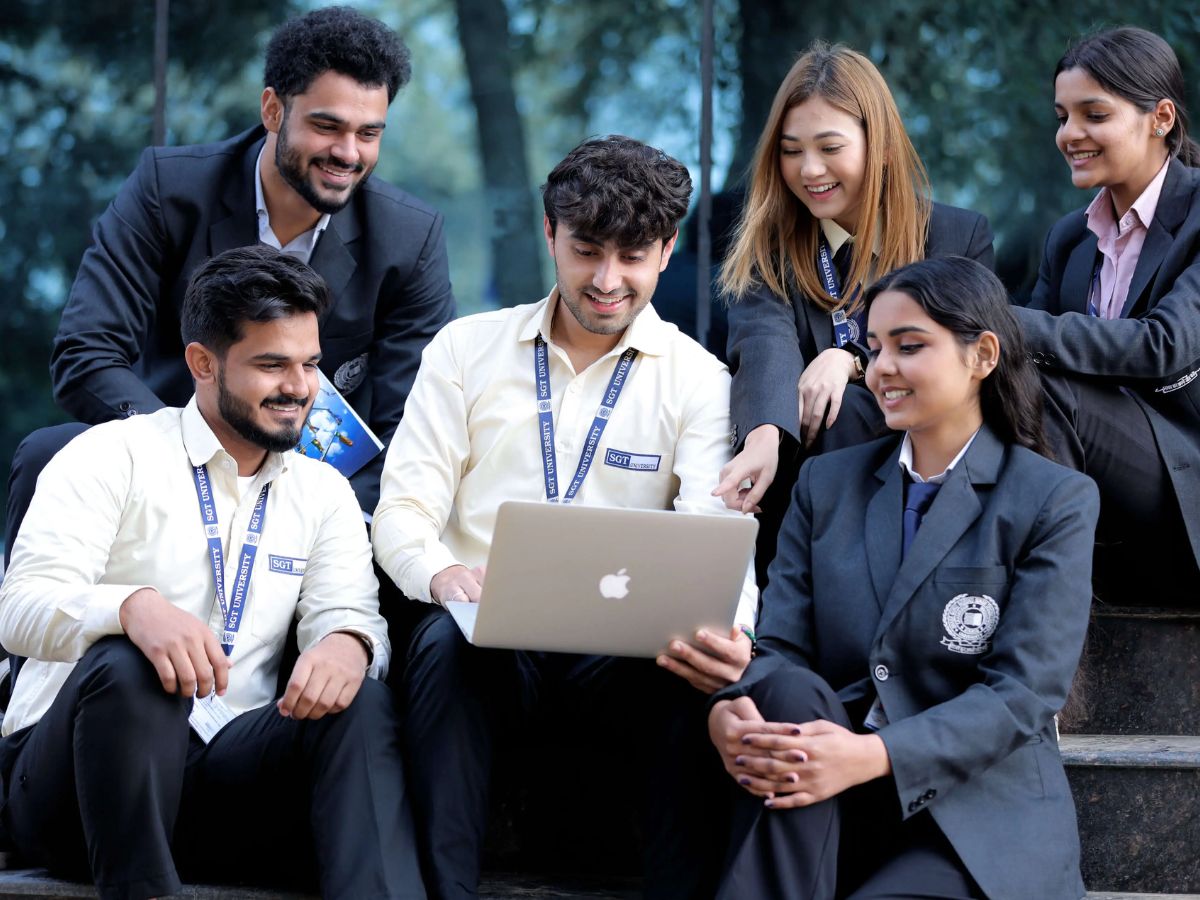 A diverse group of six college students sitting on steps outdoors. A young man holds a laptop, and everyone is smiling and looking at the screen.