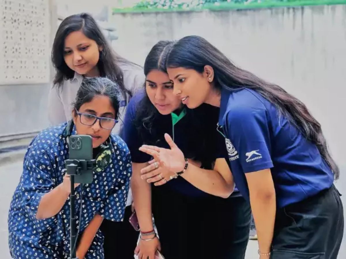 Four female students gathered around a smartphone on a tripod. They are looking at the screen and discussing a video or media project.