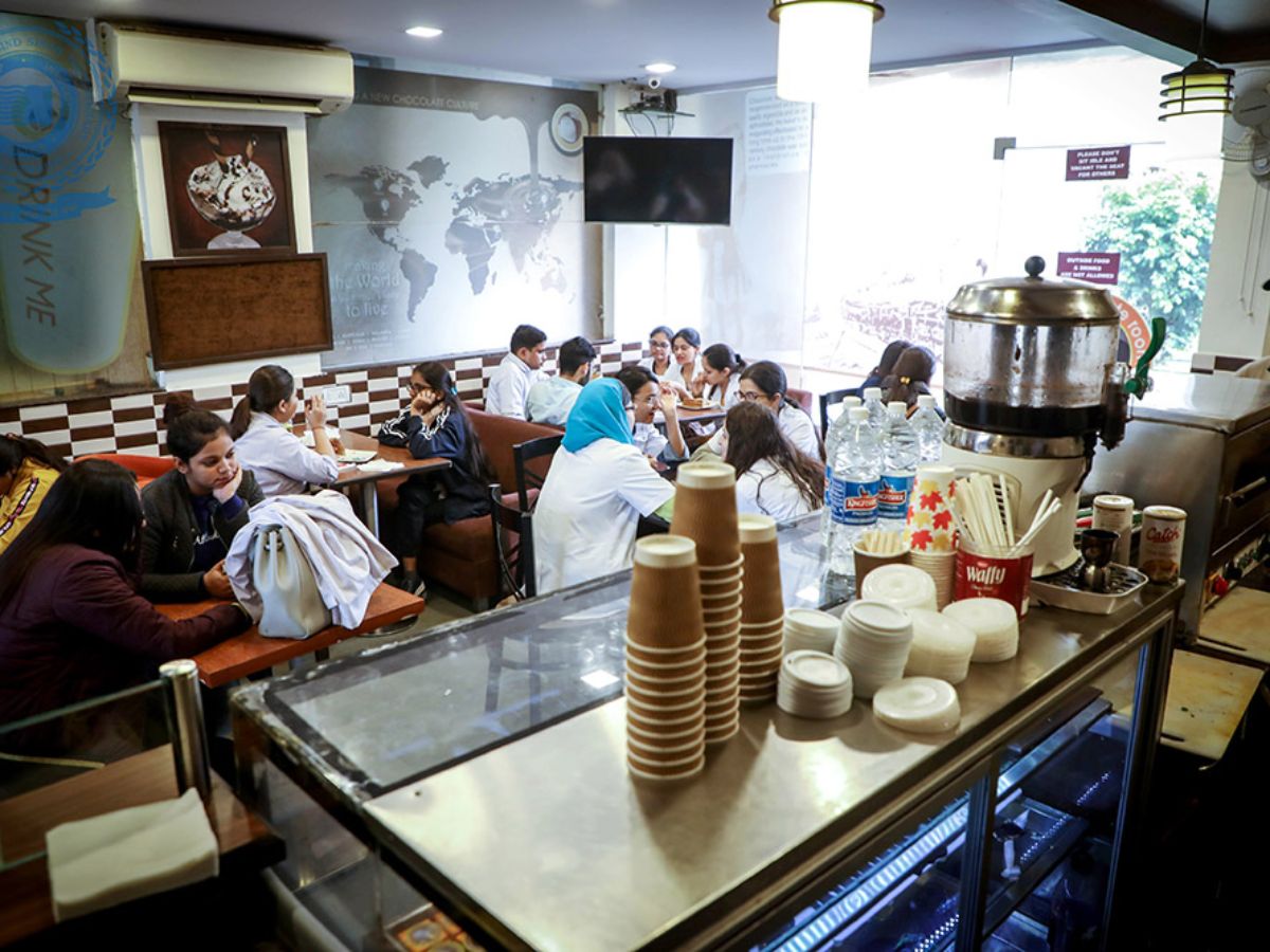 Students sit at tables inside a cafe or canteen. The foreground shows a service counter with stacks of cups, and a world map is visible on the wall in the background.