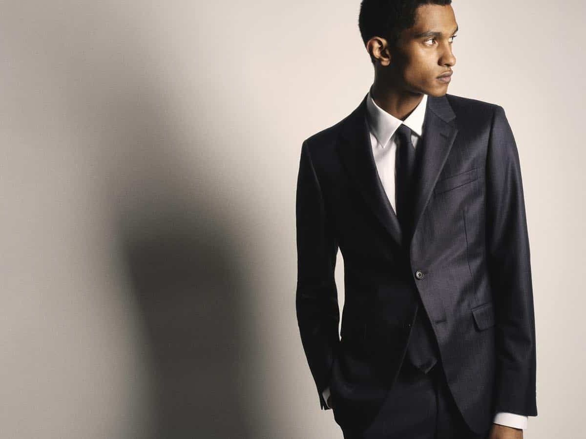 Man wearing a sleek black formal suit with a white shirt and black tie, standing against a neutral background and looking sideways with a sharp expression.