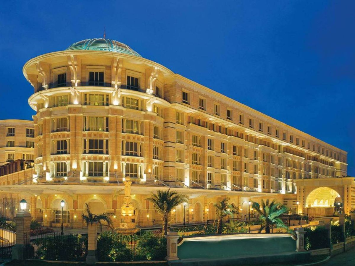 A luxurious hotel building with a large dome and traditional architecture, illuminated against a deep blue evening sky. Palm trees and landscaped gardens are visible at the entrance.
