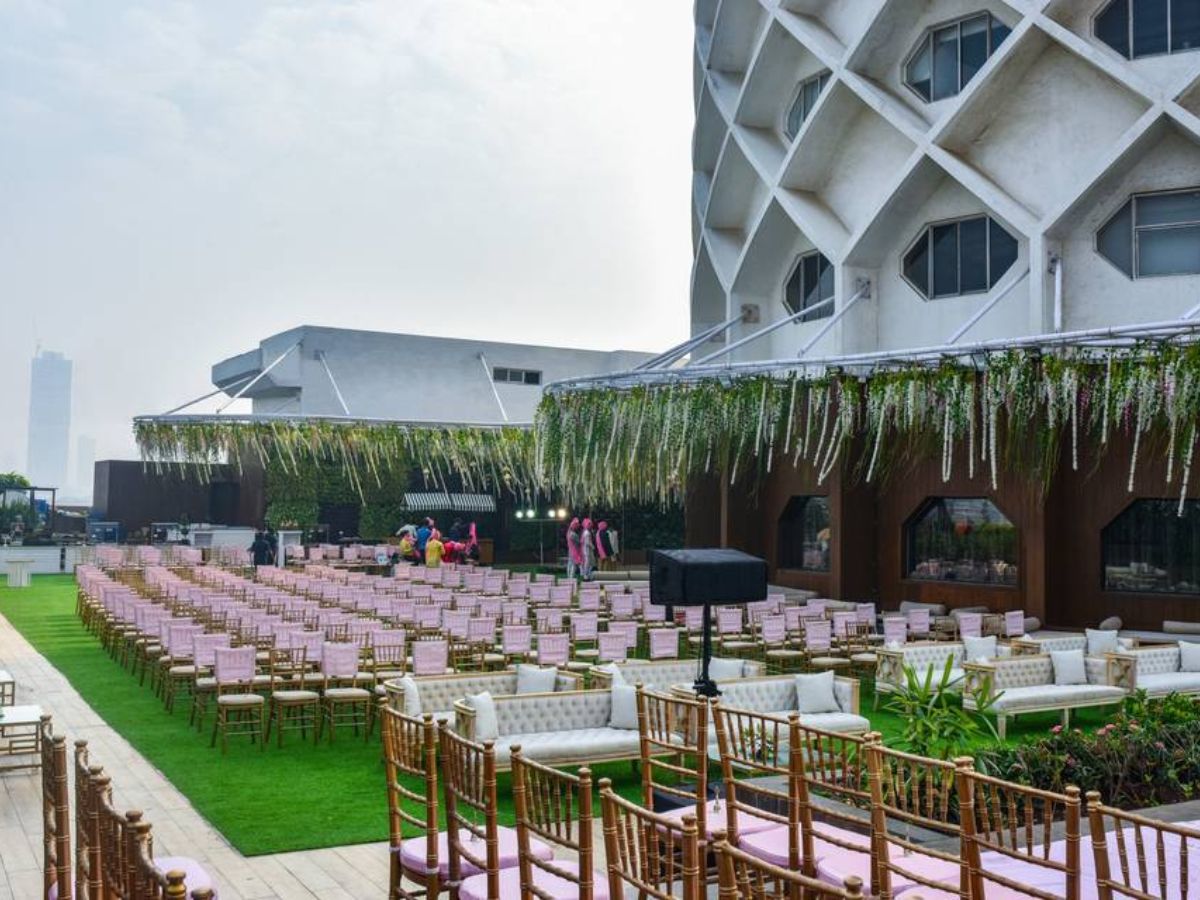 An outdoor wedding or event setup with rows of chairs with pink cushions and white sofas on a green lawn, located next to a modern building with a geometric facade. The area is decorated with hanging white floral arrangements.