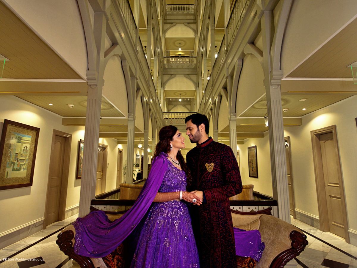 A bride and groom, dressed in traditional attire, stand facing each other in a luxurious hotel corridor with ornate arches, pillars, and an elegant, symmetrical design. The bride is wearing a sparkling purple lehenga while the groom is in a deep maroon sherwani.